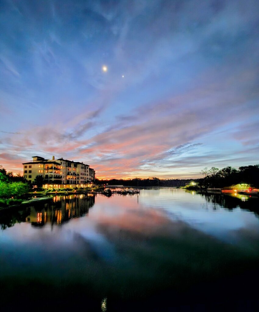 Brilliant Pre-Sunrise show across a body of water. A deep blue sky highlighted by the glow of the moon and nearby Venus is swept with thin smoke-like clouds, reflecting shades of purple and blue from the approaching sunrise at the horizon. All reflecting upon the calm, dark waters below. To the left a row of condominiums are brightly lit internally. To the right a small black dock glows from a lone street lamp.