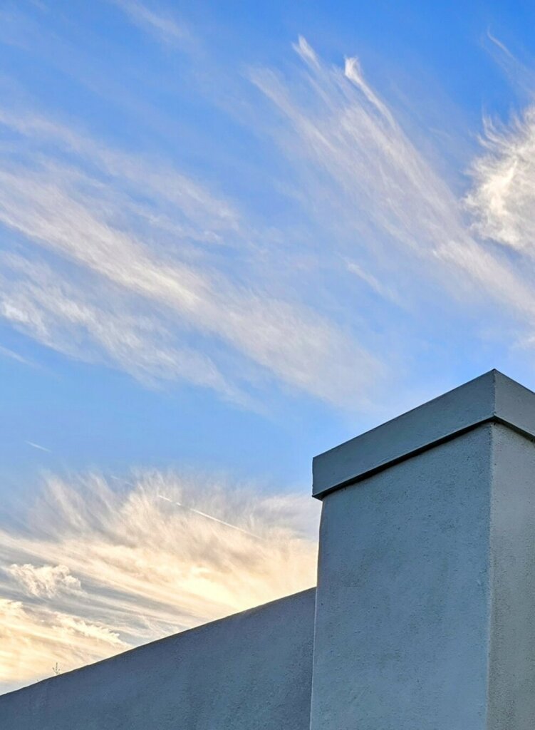 Upwards view into a bright blue sky with whispy white cloud streaks above a concrete wall.