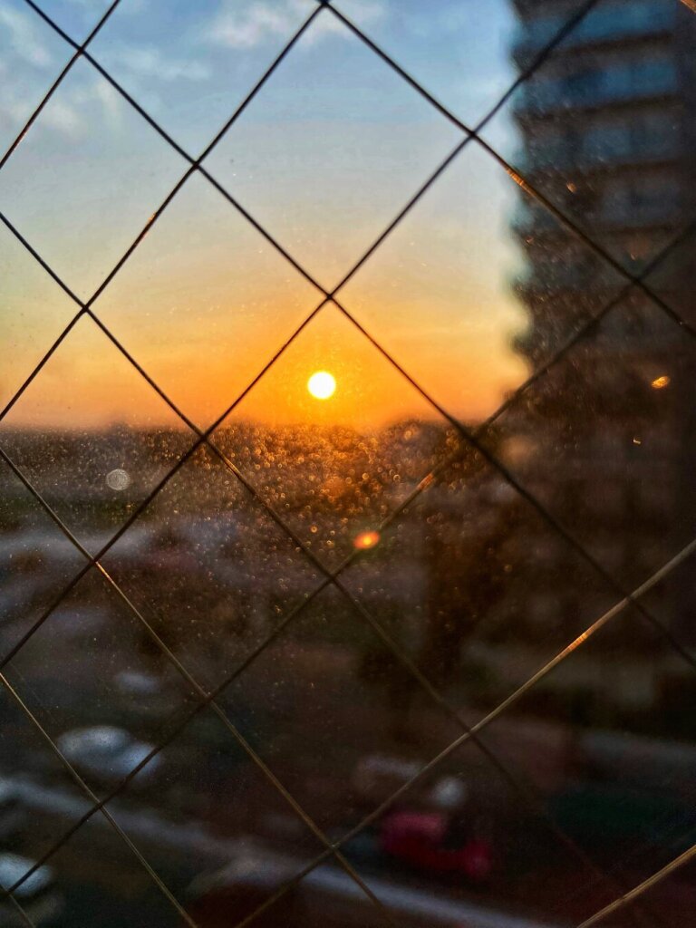 Sunset viewed through a diamond-patterned window grill with dust particles highlighted by sunlight, silhouettes of buildings visible in the background.