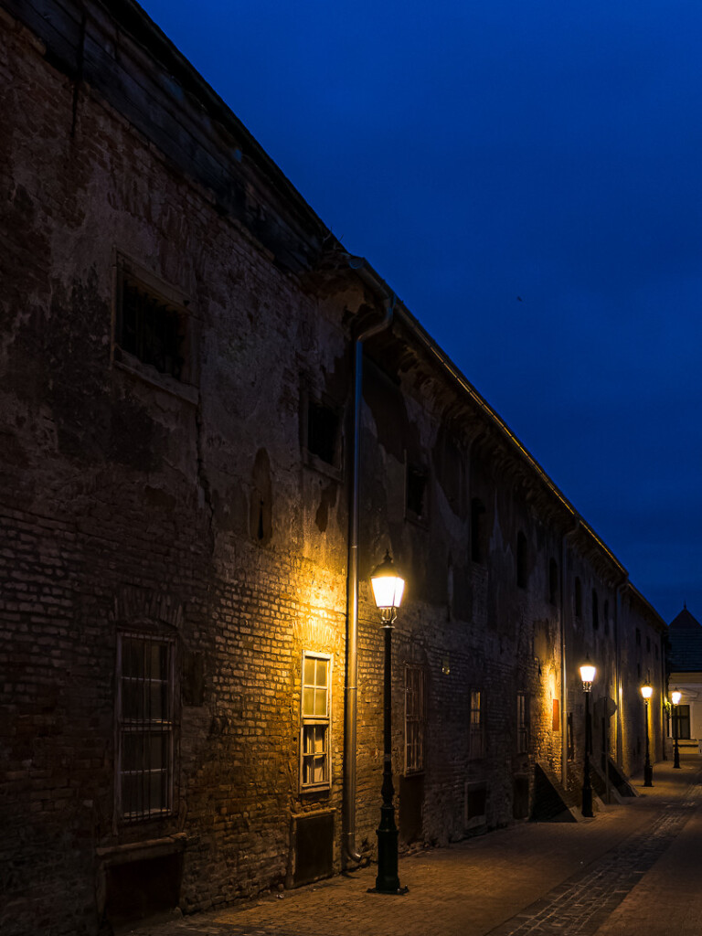 Eine Gasse mit dem Klinkerbau einer alten Brauerei am linken "Ufer". Davor, dem Bogen der Straße folgend, alte Laternen. Blaue Stunden, schönes Licht am Leuchten. 