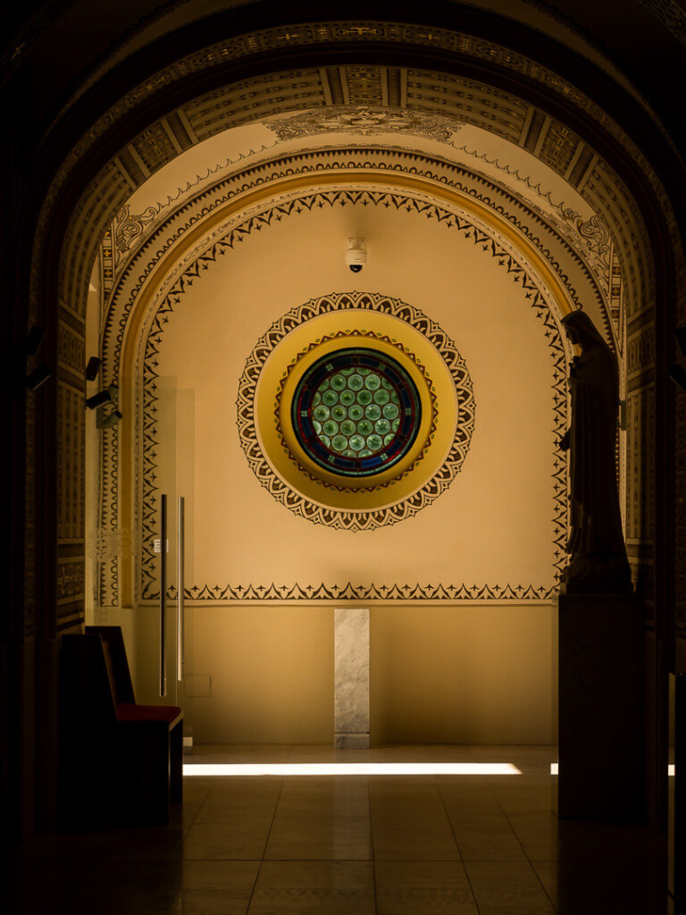 Vorraum einer Kirche, innen zur Seite fotografiert. Warmes Licht an der Wand entlang. In der Wand ein rundes "Fenster", dessen Glas aus Mosaik besteht. Davor im Dunklen Silhouetten - rechts eine Statue, links Sitzgelegenheit.