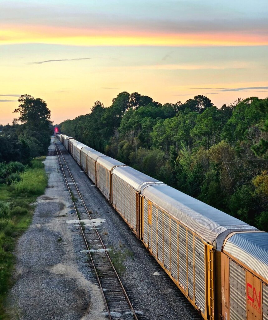 View from above of a train passing beneath a bridge, with double-decker box cars owned after the other stretching far into the distance all below the colors of an early morning sunrise with bands of pink, yellow and orange across the sky.