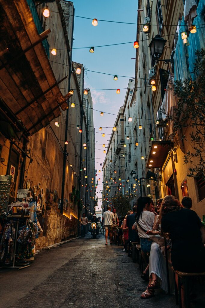 Photo of a lively street scene with people seated at tables, illuminated by charming hanging lights.