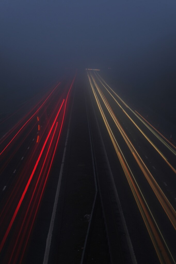 Portrait format colour image taken from a motorway bridge before sunrise, and using a 20-second exposure to create red and yellow light trails from the lights of the vehicles racing in both directions into a heavy fog that has masked any details of the surrounding landscape. On the left can be seen the red brake lights of a vehicle that is changing lanes, appearing only as three bright red dashes.