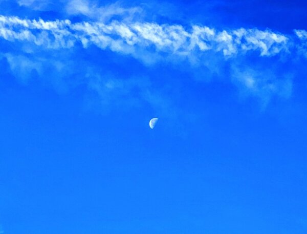 Upwards view into beautifully blue sky witha day moon visible with sweeping smoke-like clouds.