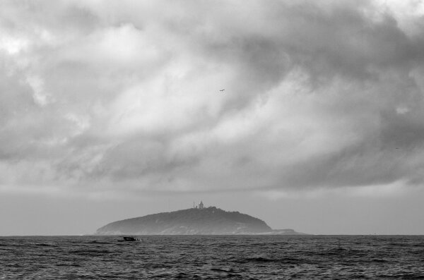 A black and white image of a distant island with a lighthouse on it. The sky is pretty cloudy and heavy.