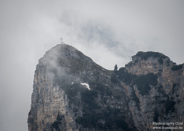 In this photograph by Franz Graf, we see a stunning mountain landscape. The dominant colors in this image are a soft shade of grey and pure white, creating a serene and peaceful atmosphere. The foreground is dominated by the image of a cross, standing proudly atop the mountain. The background is filled with a hazy fog, adding depth and dimension to the image. The sky is filled with fluffy clouds, creating a beautiful contrast against the rugged terrain of the mountain. The mountain itself is covered in trees and snow, giving the impression of a harsh yet beautiful environment. The photo is taken from a close-up perspective, showcasing the intricate details of the landscape. A person can be seen standing on the top of the mountain, adding a sense of scale and adventure to the scene. The caption for this image could be "A majestic cross stands tall on a foggy mountain, surrounded by clouds and trees, with a person standing on its peak, in a breathtaking landscape captured by Franz Graf photography."