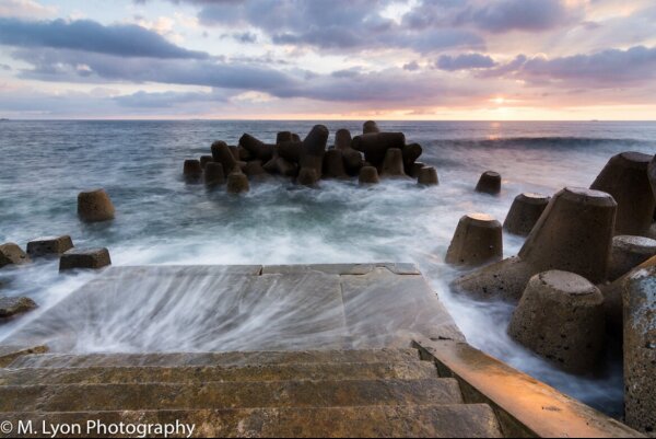 Ocean waves crashing on a concrete dock with tetrapod breakwater structures, during sunset.