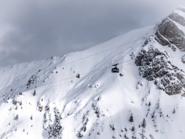 A single cable car drives from right to left through the image, with a snow covered mountain in the background. Clouds above cast sun patterns on the snow covered mountain flank.