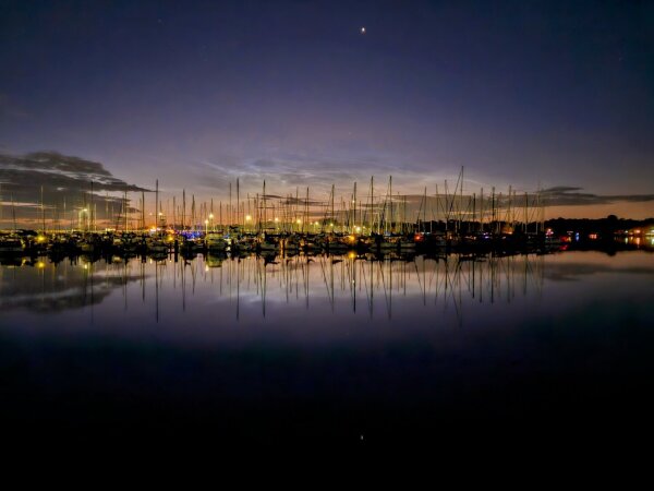 View over still, dark waters with silhouettes of sailboats and a marina with colorful lights before orange and yellow bands rising from the horizon, all casting reflections upon the water below.