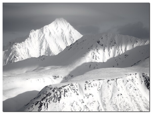 Schneebedeckte Berge. Im Hintergrund links ein mächtiger pyramidenförmiger Gipfel, die Spitze in den Wolken. Davor weitere Bergrücken, die aber nicht so schroff sind.