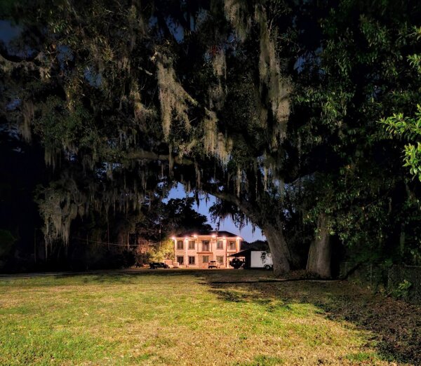 Late night view across a lush green front lawn with a large white home with pillars and bright lights making the white glow against the night sky, encircled and canopied by large oak trees draped with Spanish Moss.