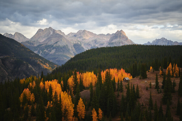 A scenic mountain landscape featuring a dense forest with vibrant autumn foliage in shades of yellow and orange. In the foreground, a small cabin sits on a hillside. The background showcases rugged, rocky mountains under a cloudy sky, creating a dramatic and picturesque view.